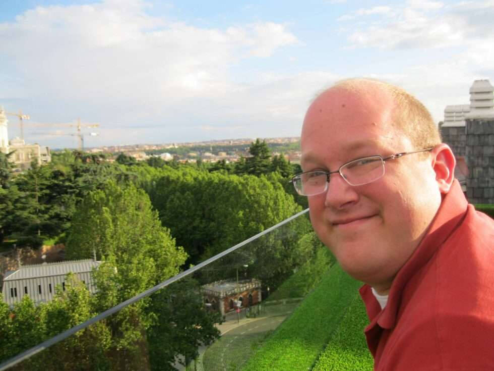 A smiling man leans over a grassy balcony rail overlooking a lush park and cityscape in bright daylight, with leafy trees and distant buildings below. He has short hair, glasses, and wears a red shirt, appearing relaxed and content in an outdoor setting.